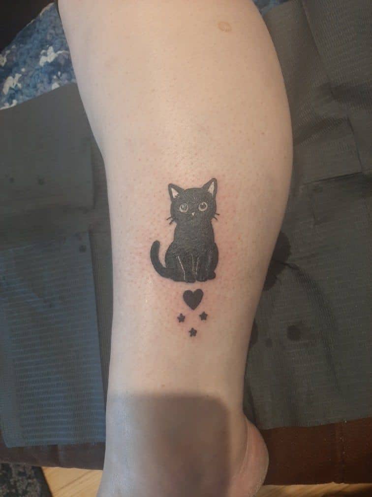 Tattoo by Jake Brewer, small black cat with a heart and stars on a lower leg