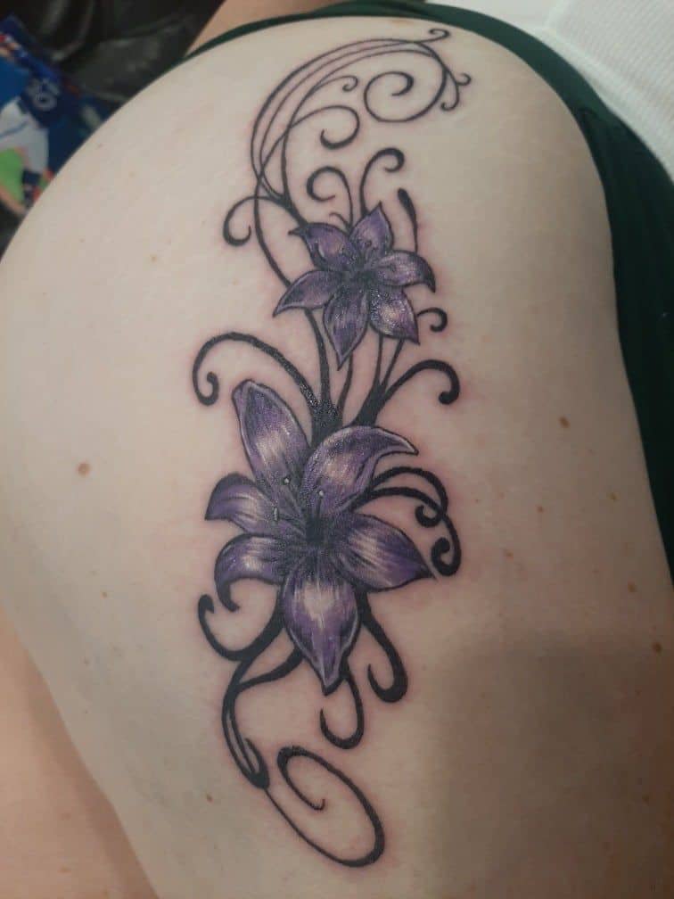 Tattoo by Jake Brewer, lilies with swirling filigree in black and grey on an upper arm