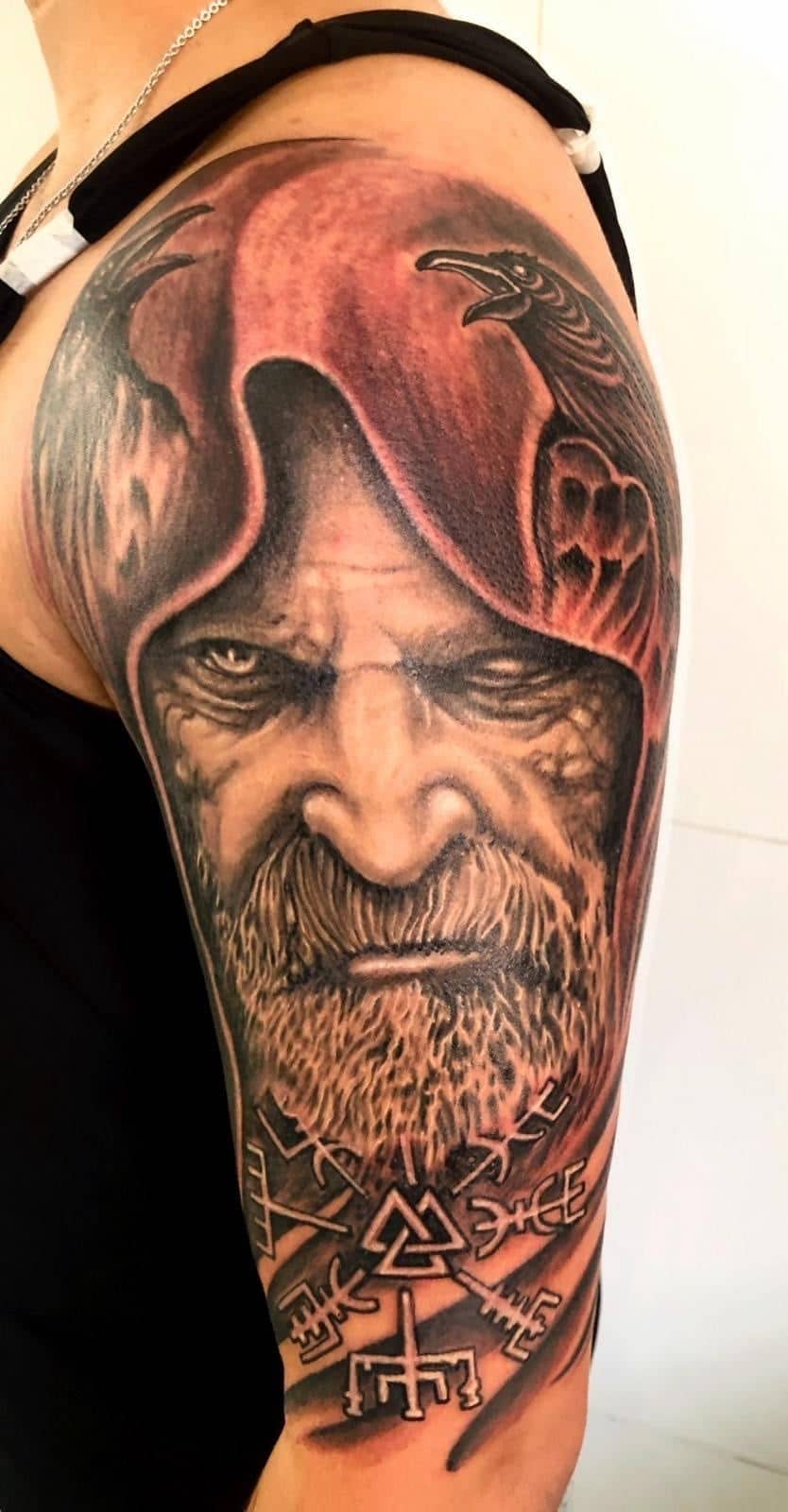 Tattoo by Ricardo Mendes, hooded bearded elder in black and grey realism with runes beneath, on an upper arm