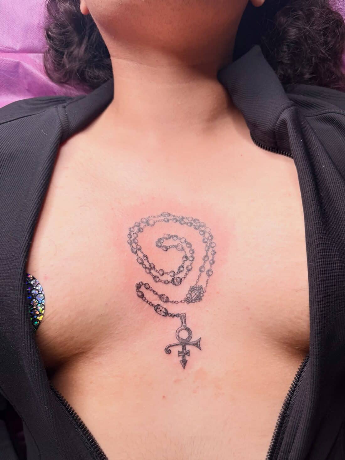 Tattoo by Apollonia Ink in Barrie, rosary with a Prince love symbol pendant on the chest